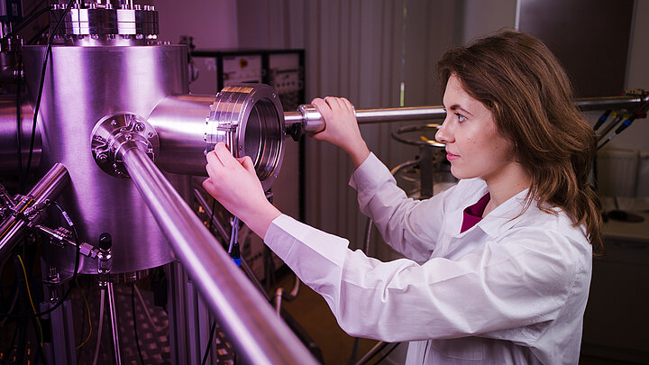Doktorantka w laboratoryjnym fartuchu stoi przed aparaturą zbudowaną z wielu metalowych rur.
